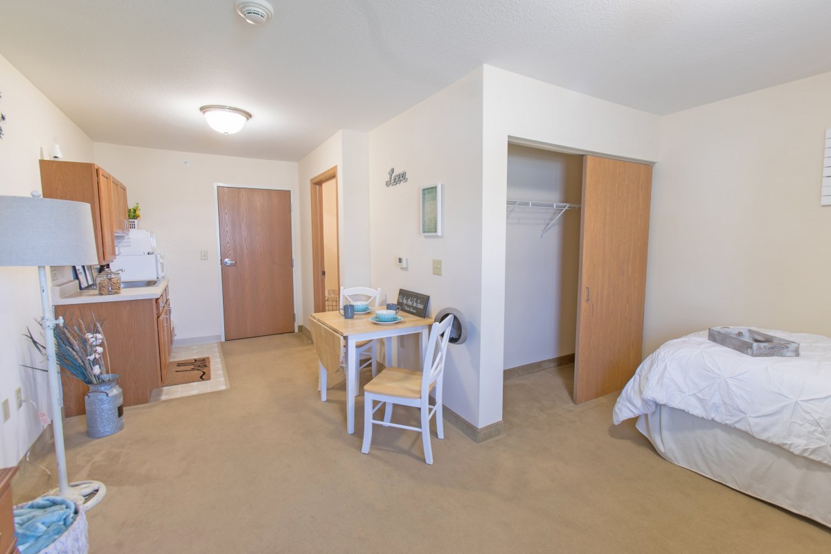 A minimalist apartment interior with a small dining table and chairs, open closet, and bed in view.