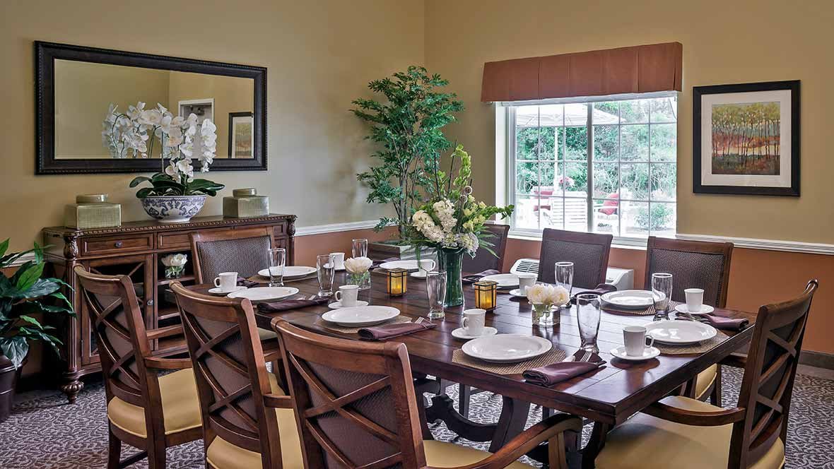 A well-decorated dining room with a large table set for a meal, surrounded by chairs and lush greenery by the window.