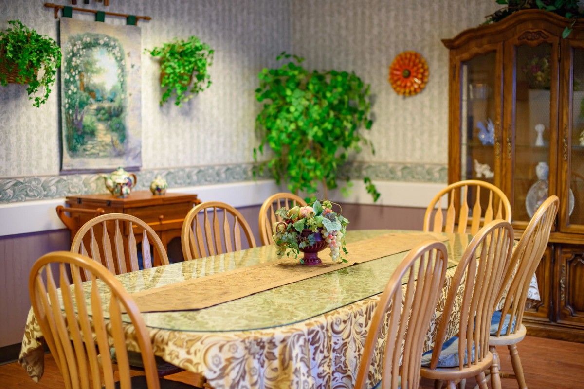 A cozy dining room featuring a wooden table and chairs, adorned with a floral tablecloth and surrounded by lush indoor plants and wall art.