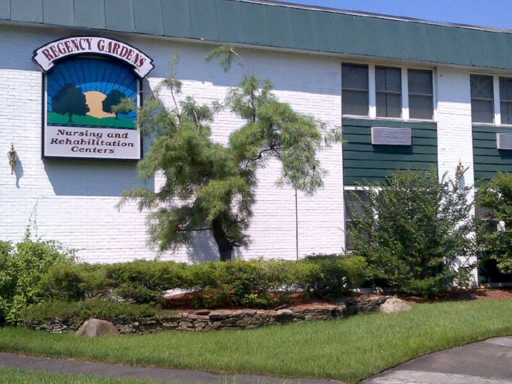 A white school building with a green and blue sign, surrounded by greenery.