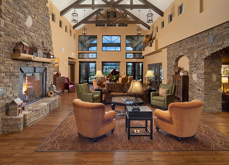 A spacious and rustic living room with a high ceiling, featuring wood beams, a stone fireplace, cozy seating, and large windows.