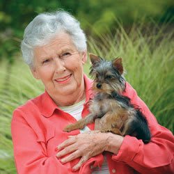 An elderly woman smiling and holding a small dog outdoors.