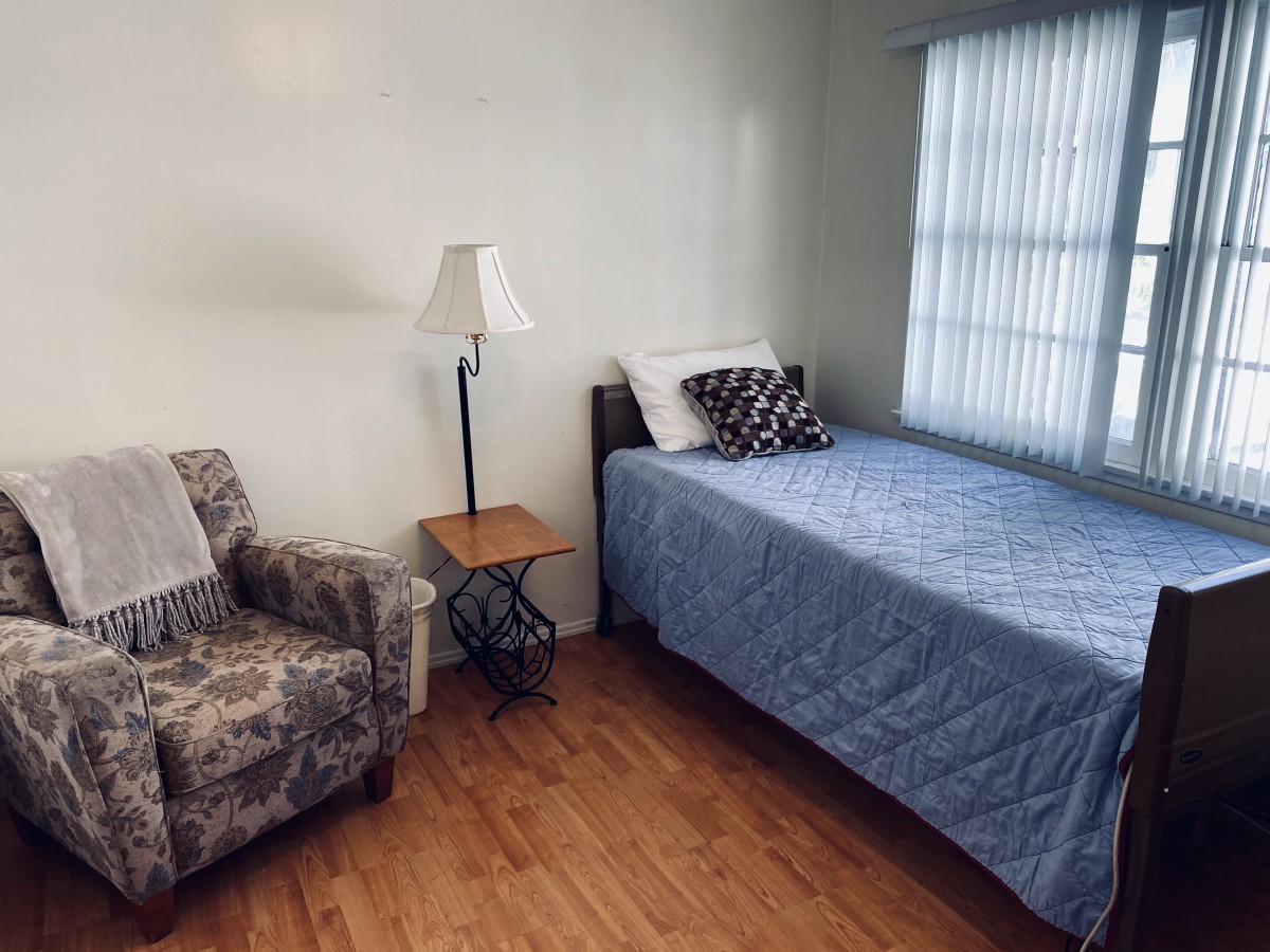 A minimalist bedroom with a single bed, a patterned chair, and a standing lamp beside a window.