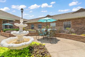 A sunny courtyard with an elegant fountain and outdoor seating under a blue umbrella, surrounded by a brick building.