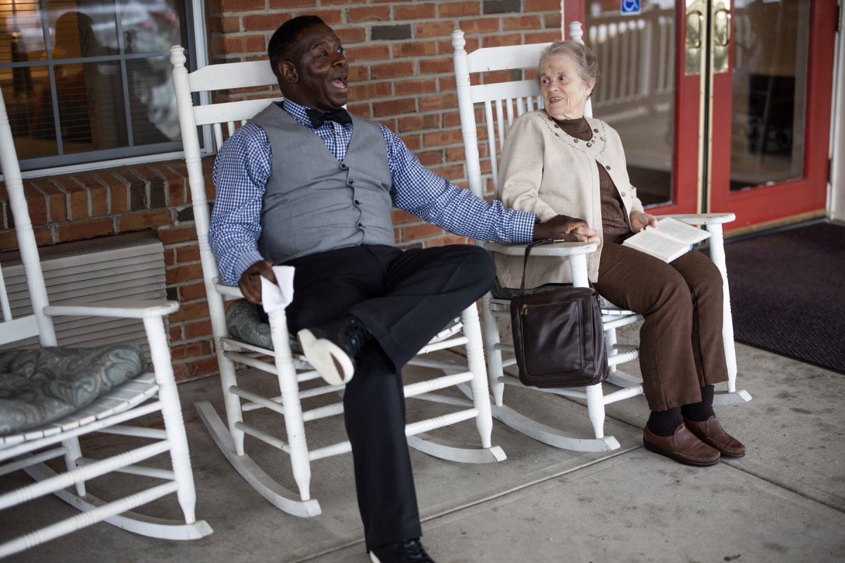 An elderly couple enjoys a relaxed conversation while sitting on rocking chairs outside.