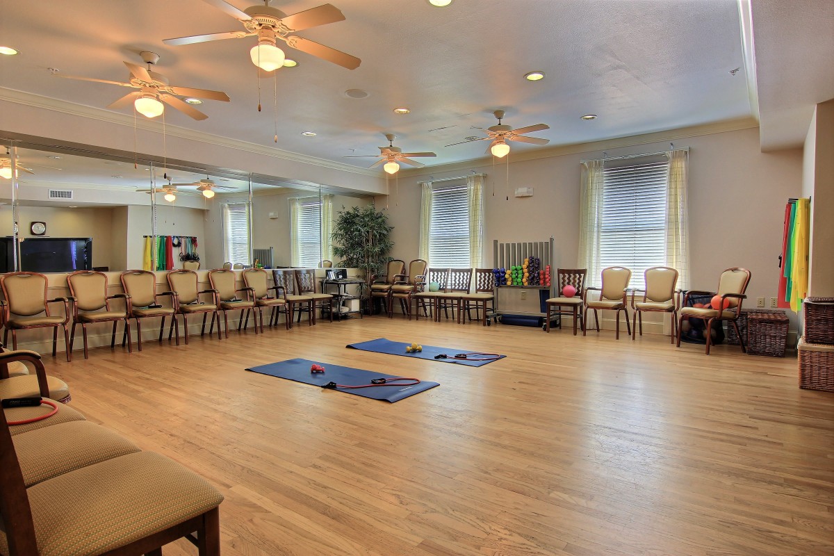 A spacious exercise room with wooden flooring, yoga mats, and chairs arranged along the walls, illuminated by natural light from large windows.