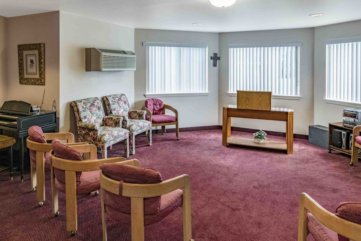 A small chapel with chairs arranged around a pulpit, featuring a cross on the wall and carpeted flooring.