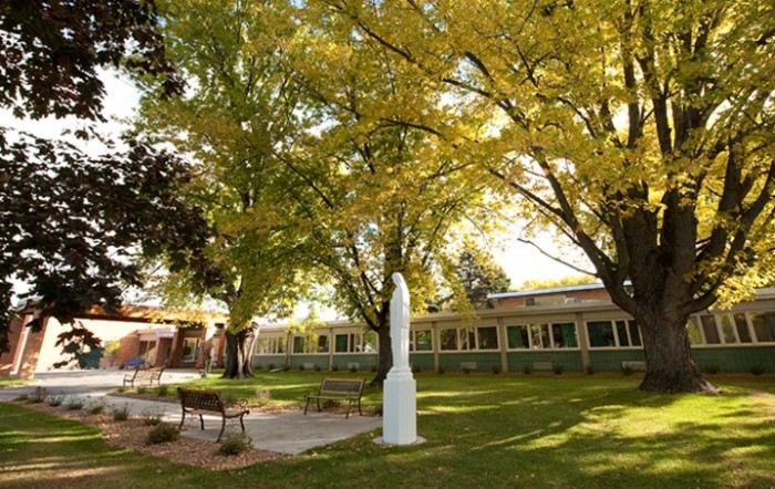 A peaceful garden area with a white statue, surrounded by trees and benches, adjacent to a building.