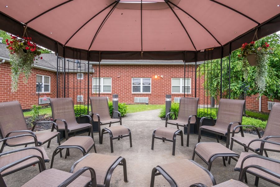 An inviting gazebo with comfortable seating surrounded by a garden and brick buildings.