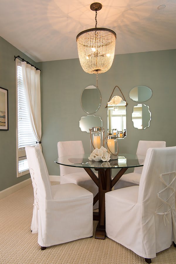 A modern dining room featuring a round glass table with white upholstered chairs, decorative mirrors on the wall, and soft ambient lighting.