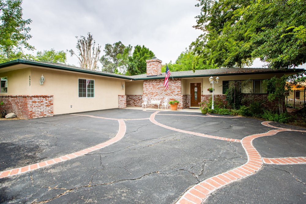 A suburban home with a spacious driveway lined by brick detailing, surrounded by trees and an American flag near the entrance.