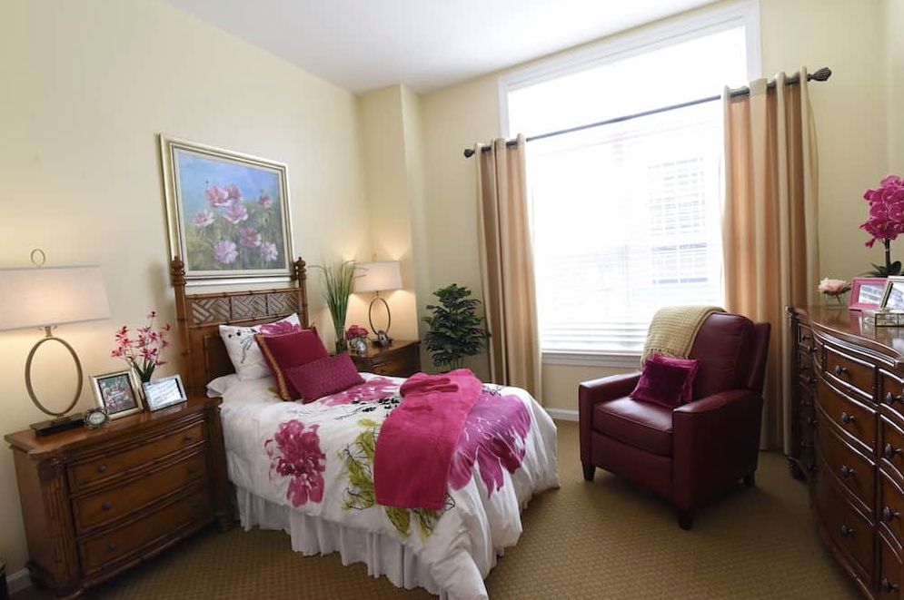 A cozy bedroom featuring a single bed with floral bedding, a comfortable armchair, and decorative elements including a painting and curtains.