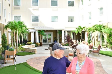An elderly couple stands in front of a building with a garden and palm trees.