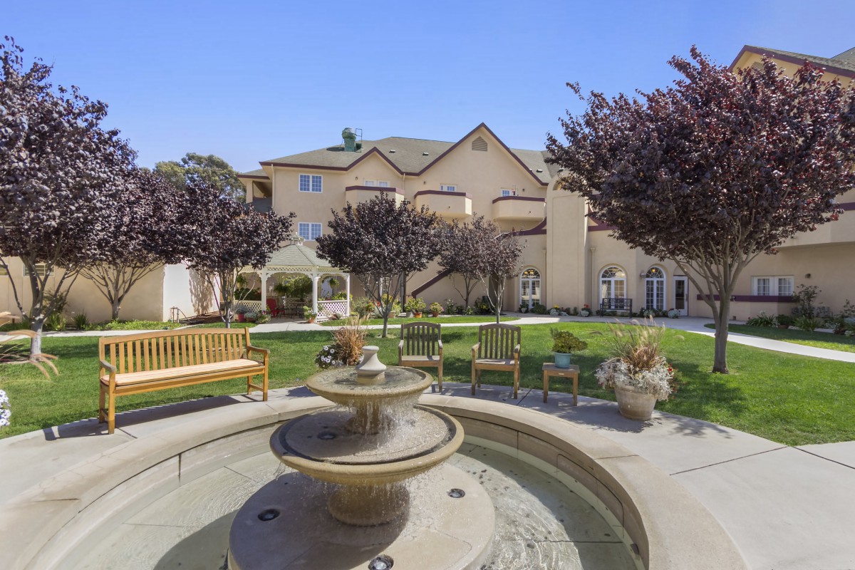 A peaceful courtyard with a fountain, surrounded by benches and landscaping in front of a building.