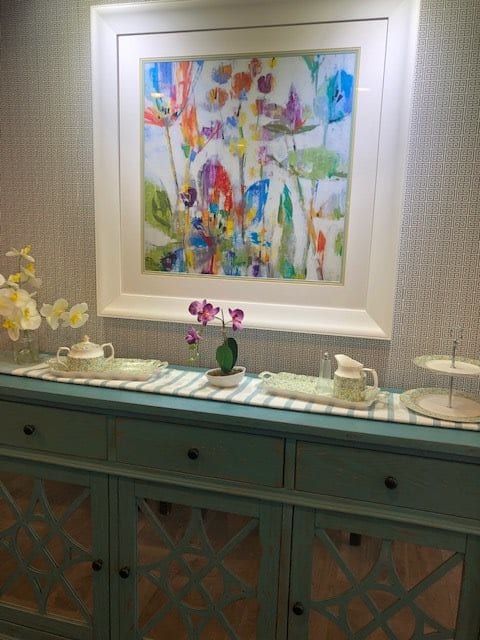 A decorated sideboard with teaware, flowers, and a colorful painting above.