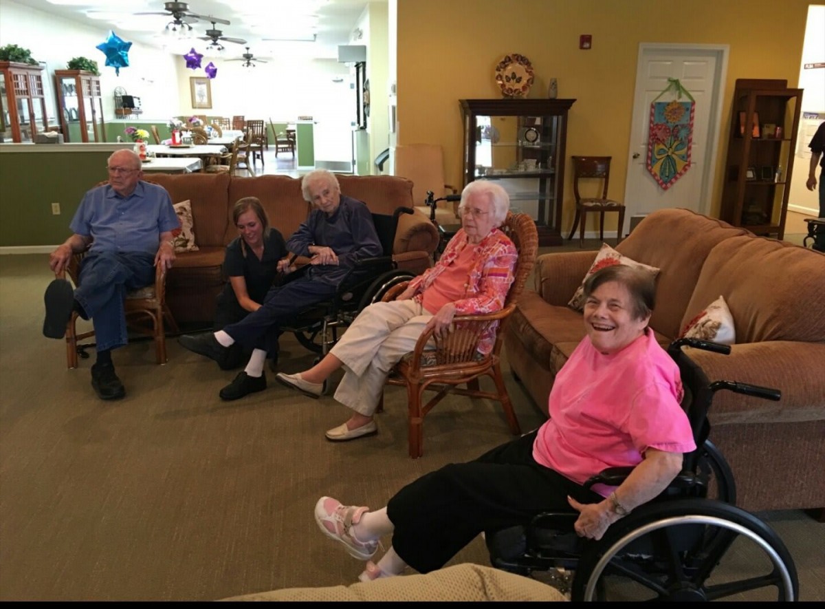 A group of elderly people gathered in a living room, one person is seated in a wheelchair.