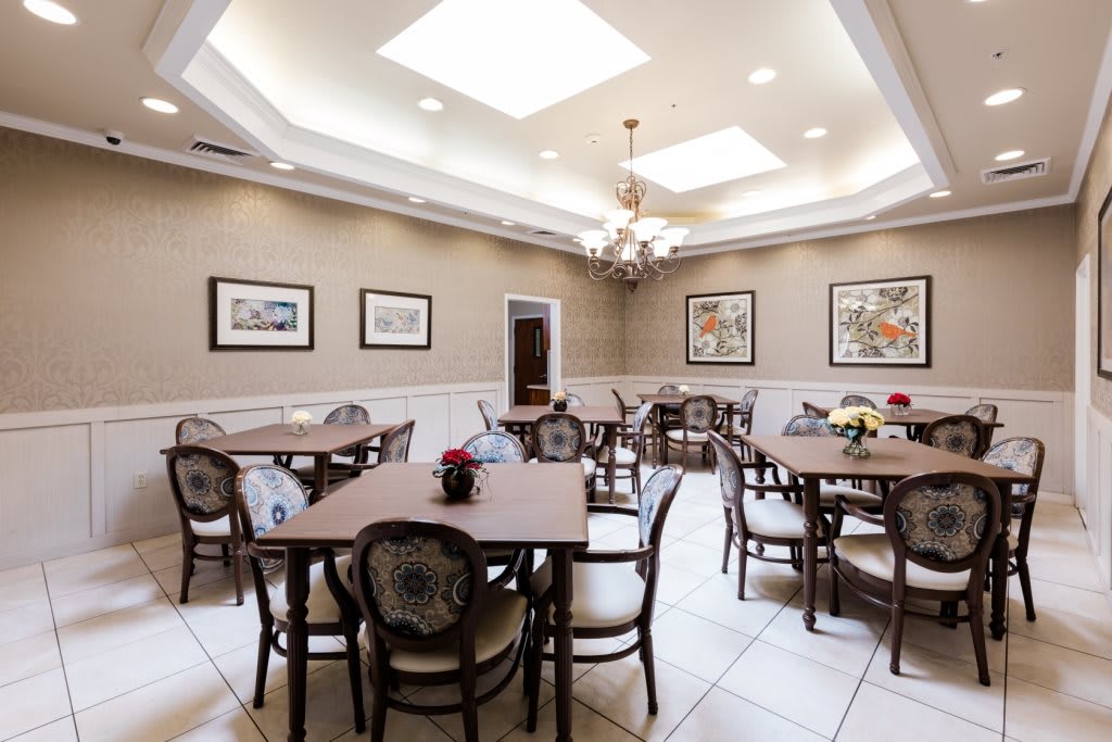 A well-lit dining room featuring multiple tables and chairs, decorated with framed artwork on the walls and a central chandelier under skylights.