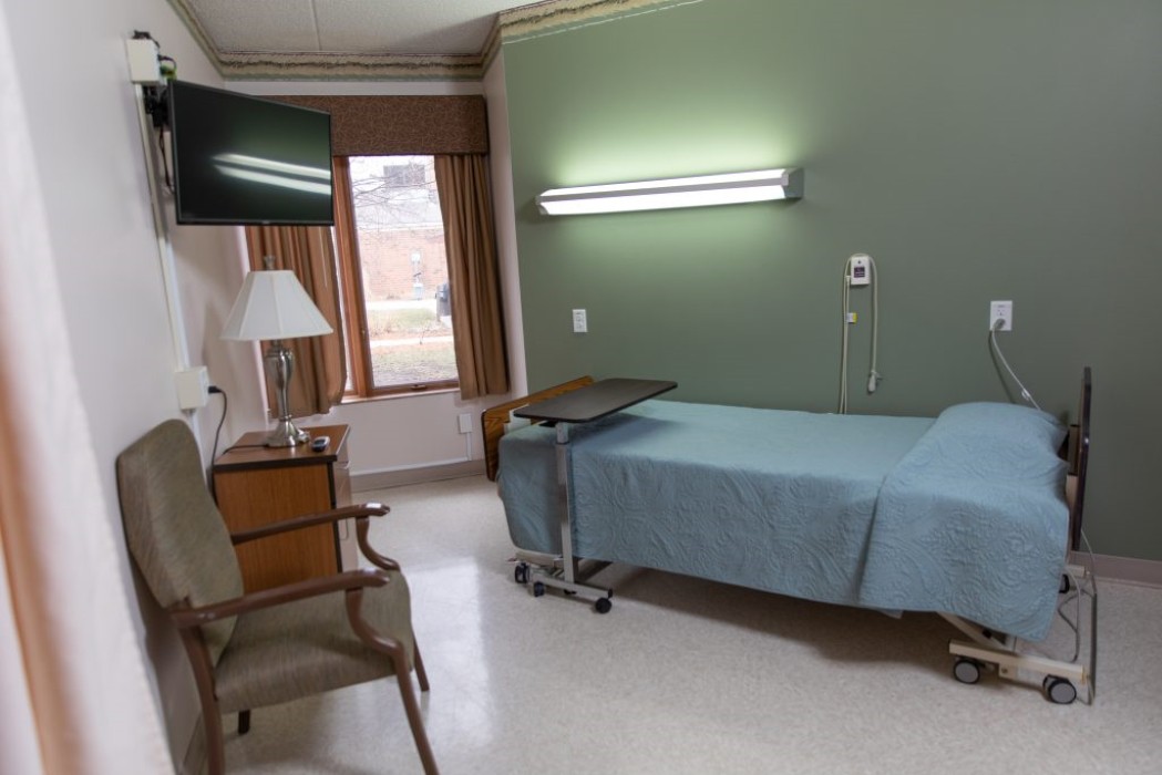 A hospital room with a single bed, television, chair, and window with curtains.
