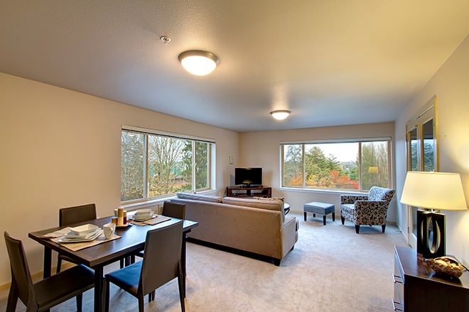 A spacious, well-lit living room with a cozy seating arrangement and a dining table, featuring large windows that bring in natural light.