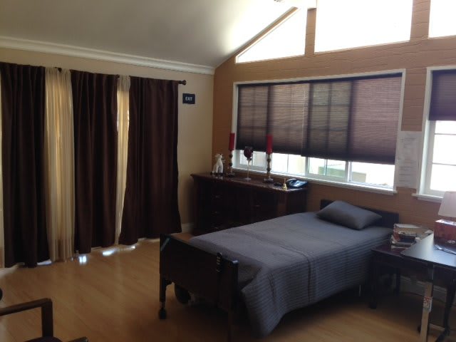 A cozy bedroom with a single bed, dark curtains, and sunlight filtering through the window blinds.