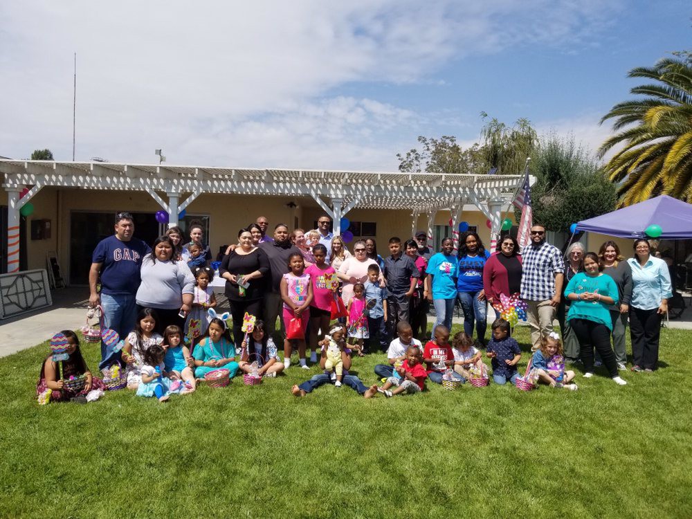 A large group of people, including adults and children, are gathered outdoors on a sunny day for a family celebration.