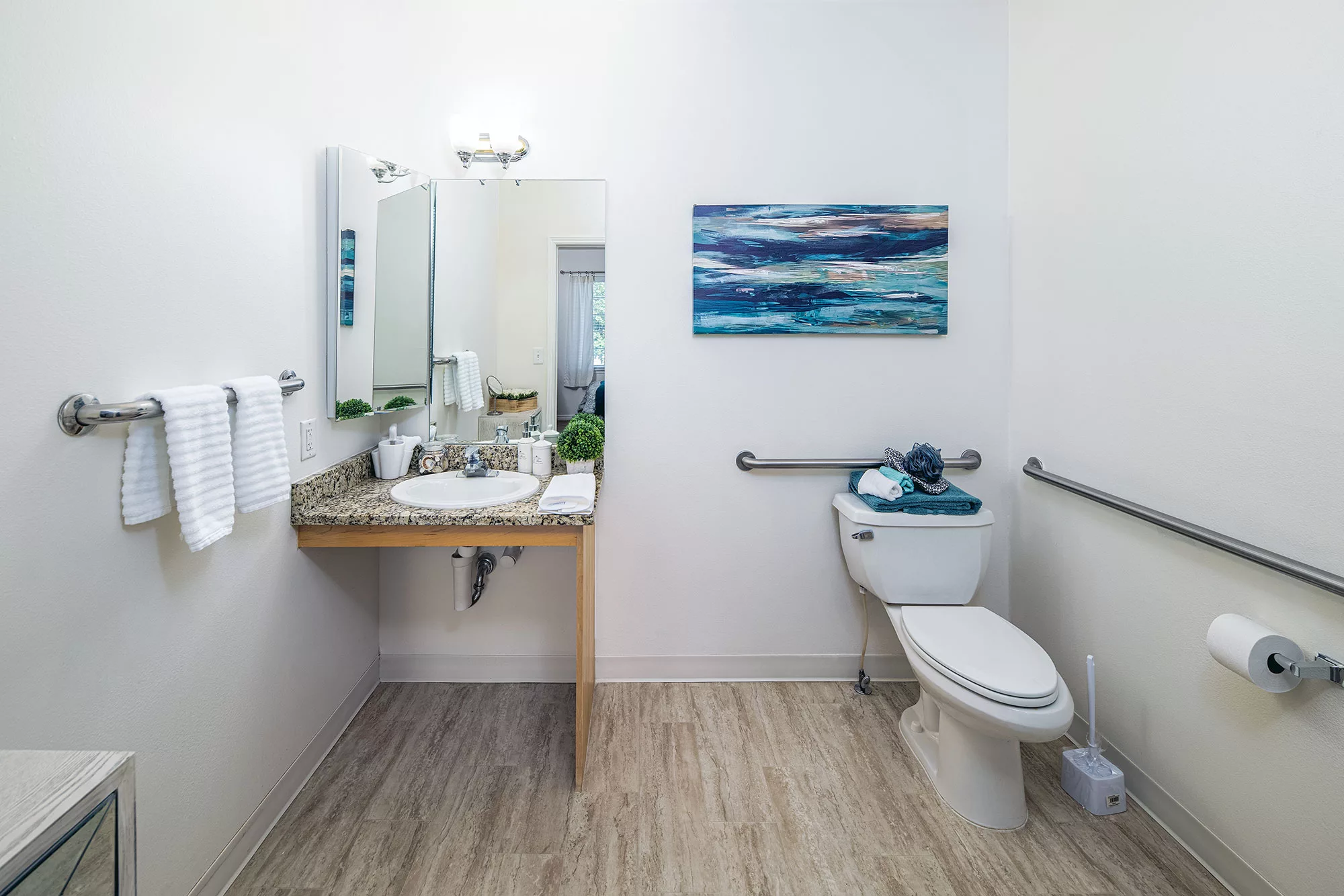 A modern bathroom with a sink, mirror, and toilet featuring handrails, and a wall-mounted painting.
