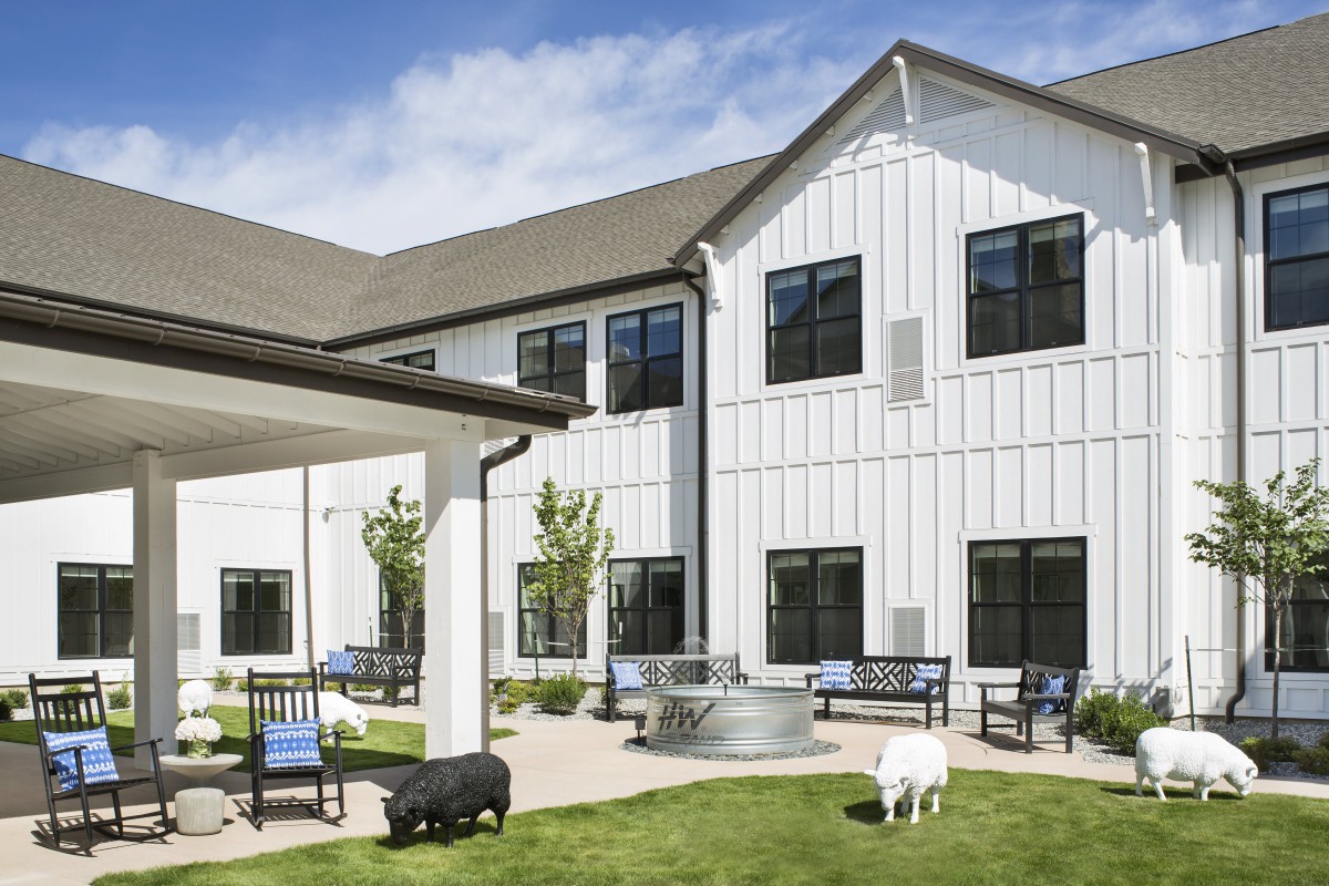 A two-story building with a neatly maintained yard featuring benches, a small lawn area, and two animals roaming freely.