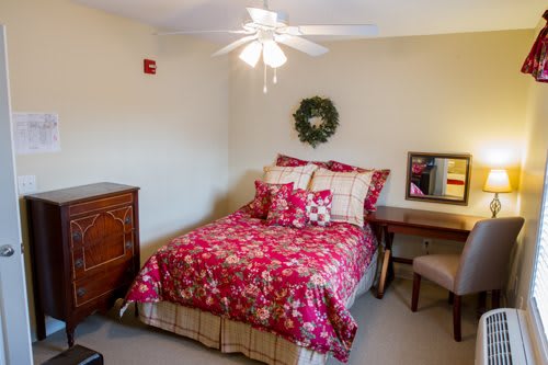 A cozy bedroom with floral red bedding and a ceiling fan, featuring a dresser, desk, and chair.