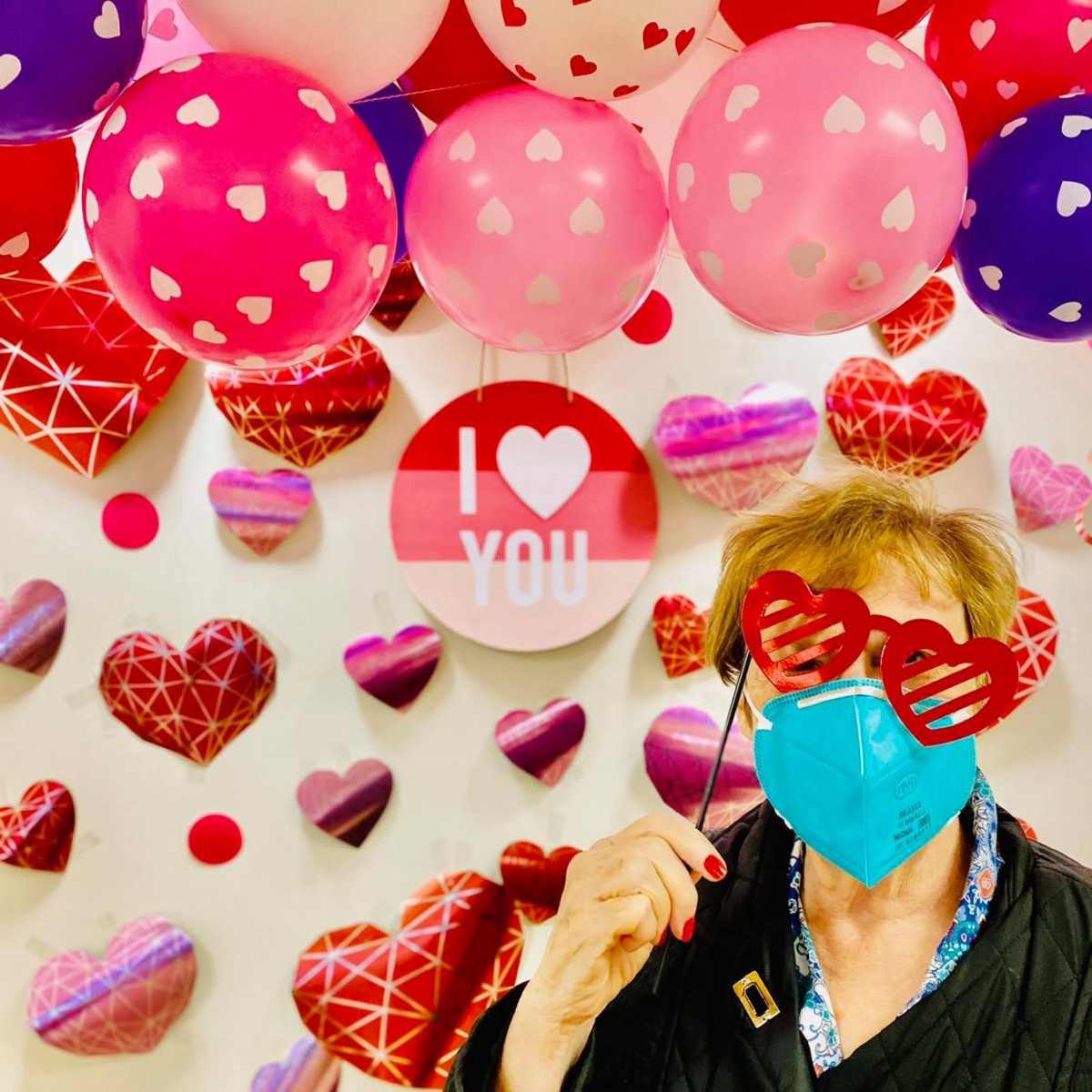 A festive Valentine's Day scene with heart-shaped decorations, balloons, and a person wearing a mask and novelty glasses.