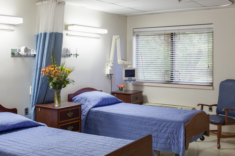 A clean and well-organized hospital room with two beds, bedside tables, and a vase of flowers near the window.