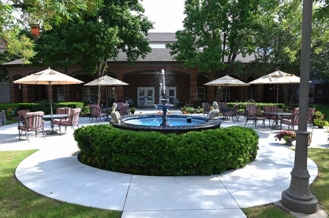 A picturesque outdoor patio featuring a central fountain surrounded by lush greenery and tables with umbrellas.