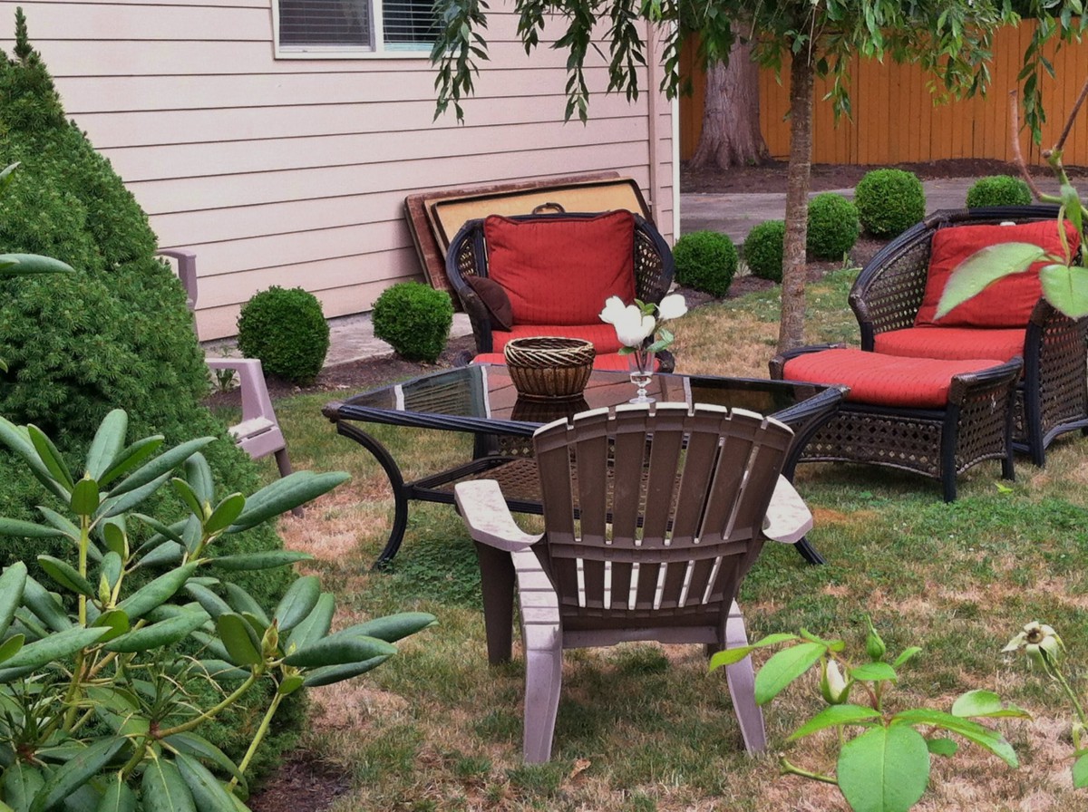 A cozy backyard setup featuring patio furniture with red cushions, a glass coffee table, and surrounding greenery.