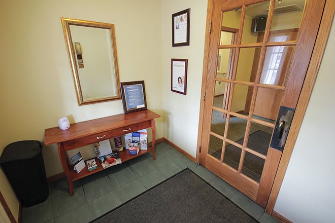 A cozy entryway with a wooden table, mirror, certificates on the wall, and a glass-paneled door.