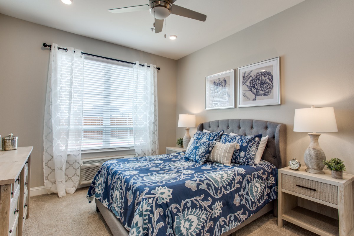 A cozy bedroom featuring a bed with blue patterned bedding, artwork above the headboard, and natural light coming through a window with white curtains.