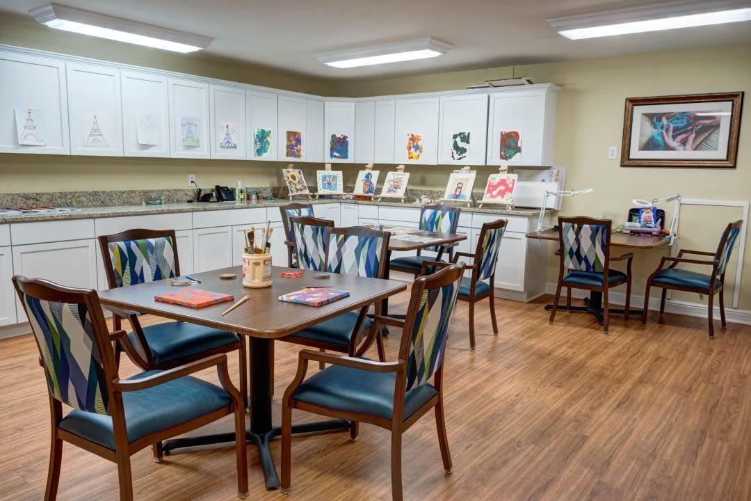 A cozy room with multiple tables and chairs, decorated with artwork and equipped with kitchen cabinets.