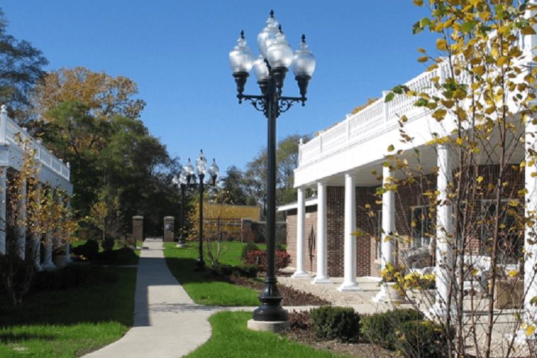A scenic outdoor view featuring a decorative lamp post along a pathway, adjacent to a building with columns and surrounded by grass and trees on a sunny day.