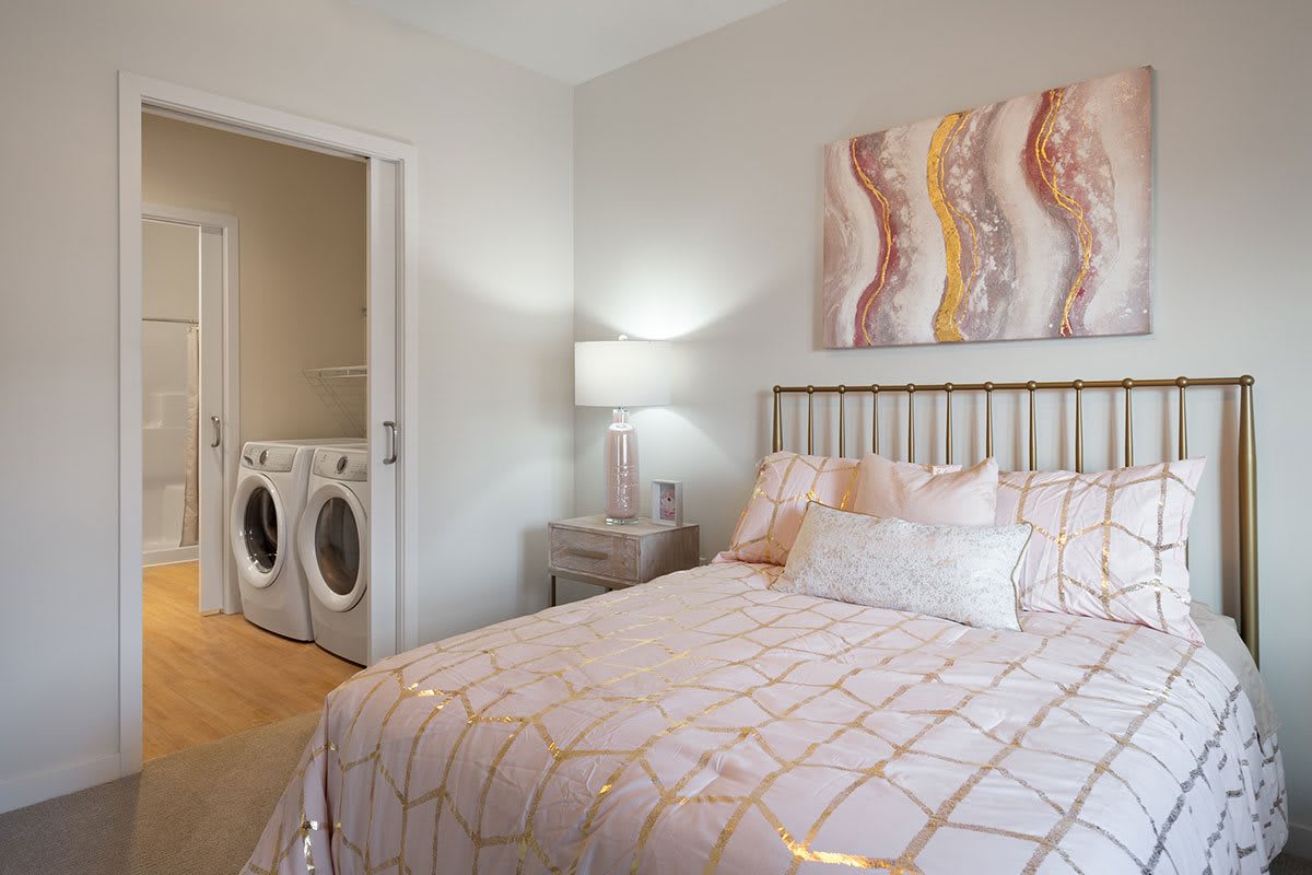 A cozy bedroom with a laundry area visible, featuring a bed, lamp, and artwork on the wall.