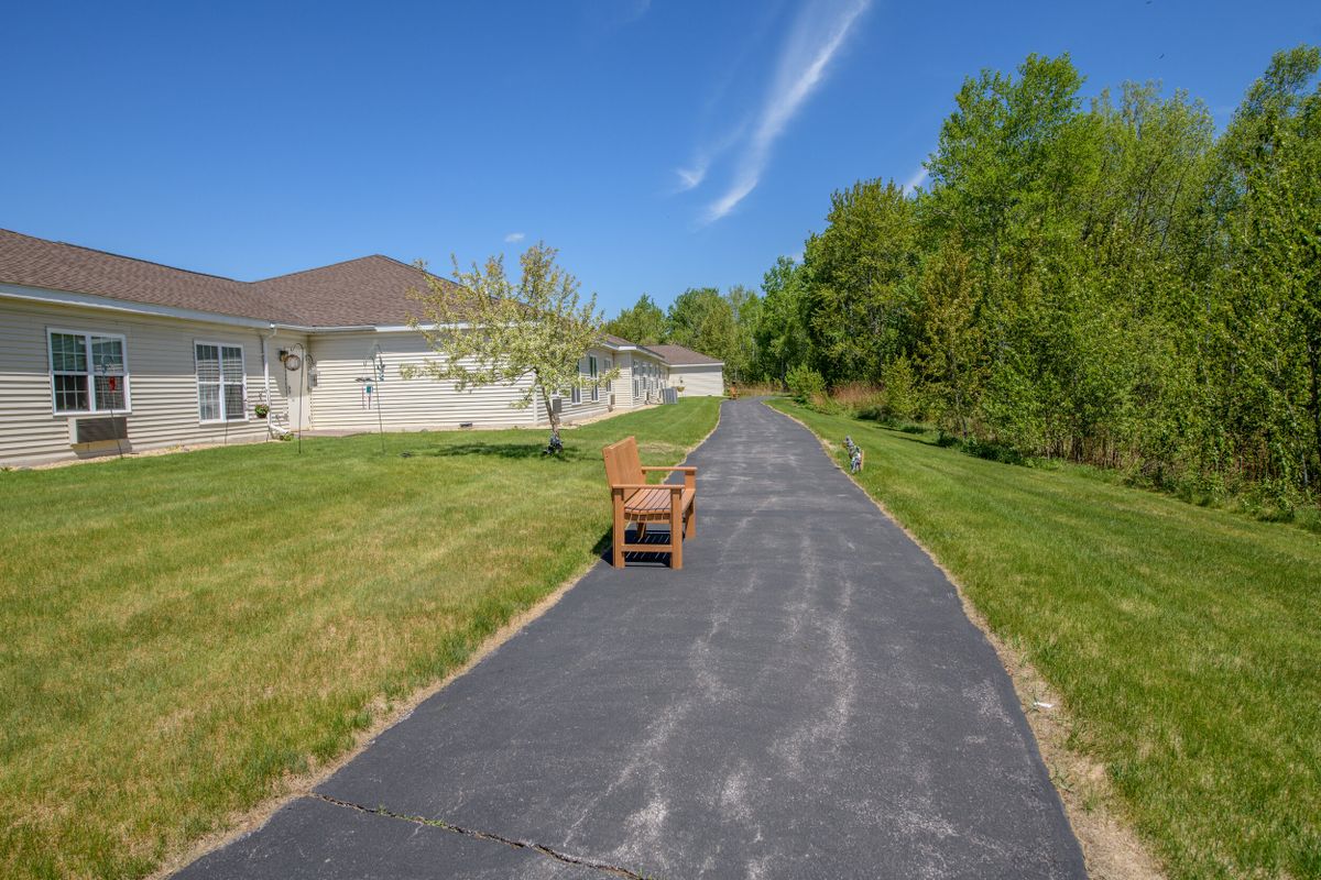 A wooden chair sits in the middle of a paved driveway leading to a house surrounded by trees and clear blue sky.