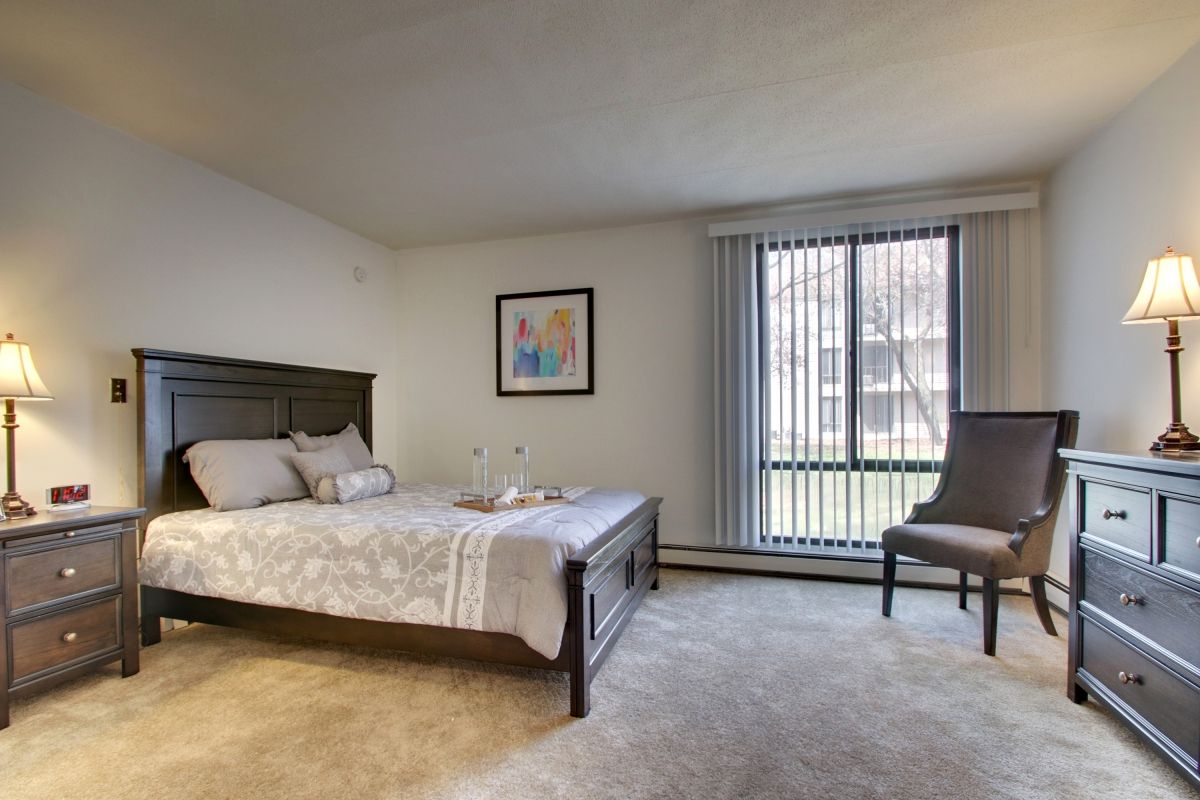 A minimalist bedroom with a large bed, decorated in neutral colors and featuring a window with vertical blinds.