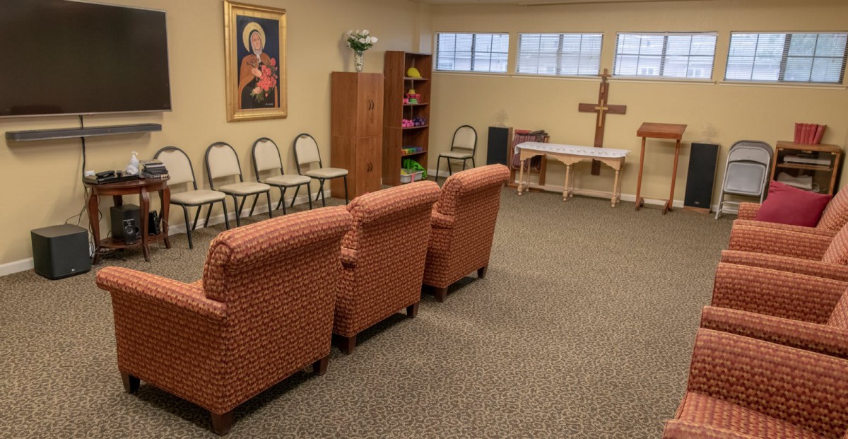 A cozy, carpeted room with chairs arranged around a cross and a religious painting on the wall.