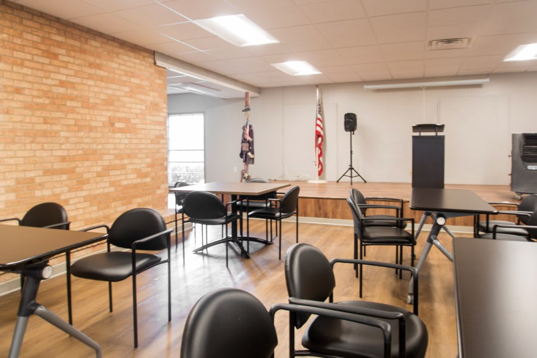An empty meeting room with chairs arranged around tables, a podium, and flags in the background.