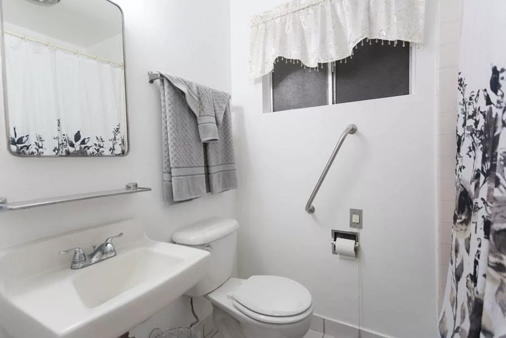 A clean, white bathroom with a sink, mirror, towels, and a grab bar next to the toilet.