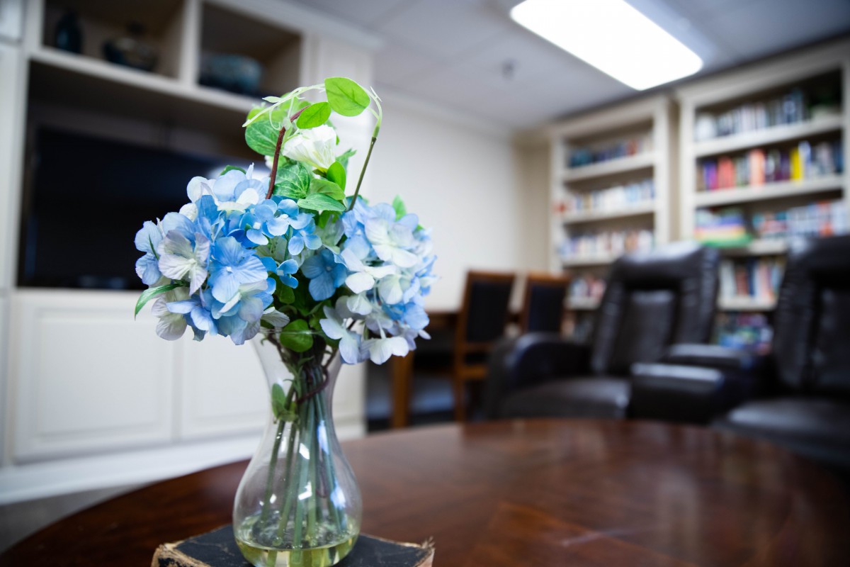 A cozy office setting with a vase of blue and white flowers on a wooden table, surrounded by bookshelves and chairs.