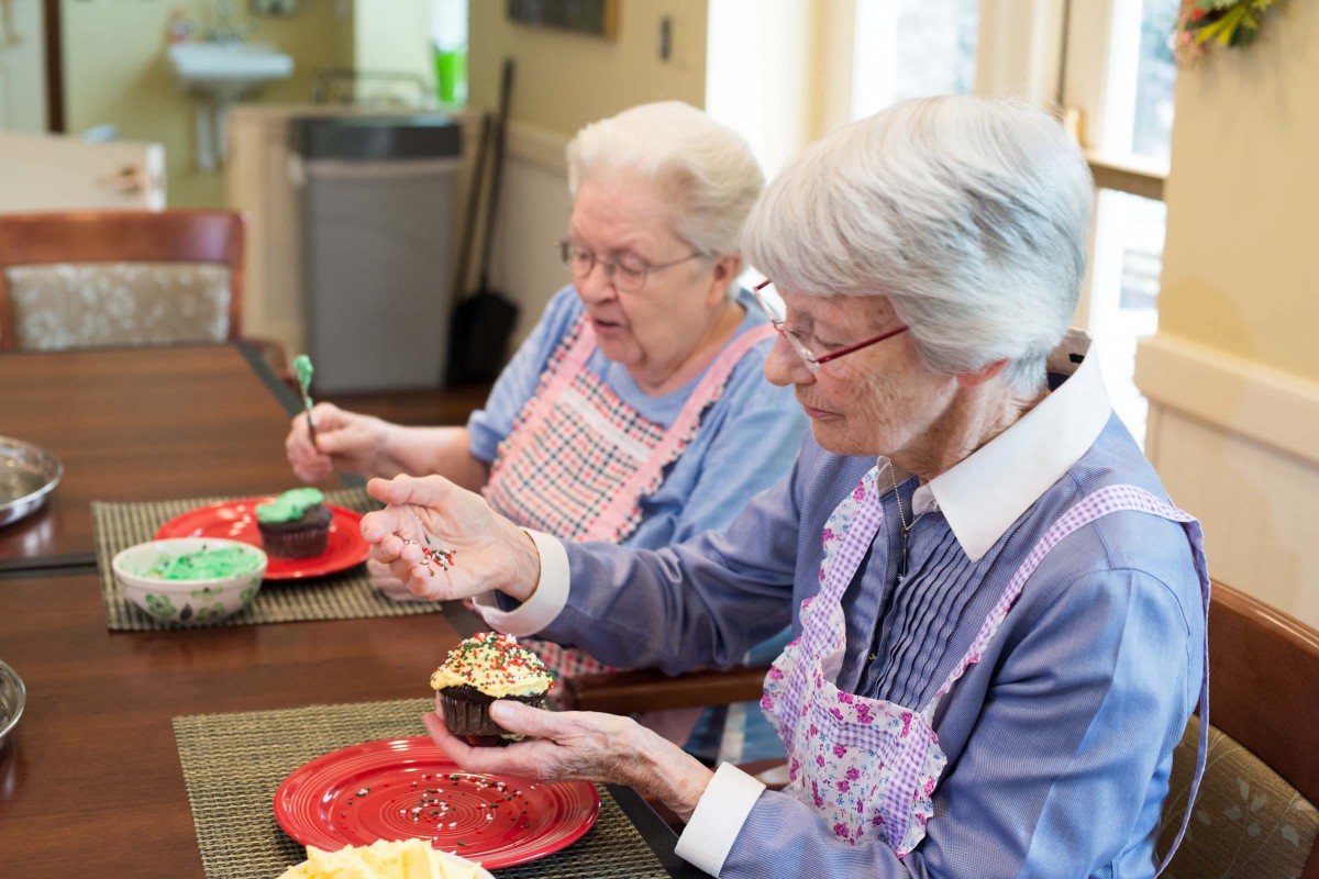 Two elderly women enjoying decorating cupcakes in the kitchen.