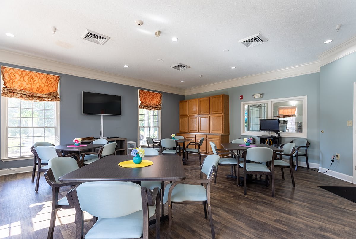 A well-lit community room with several tables and chairs, a television, and decorative elements.