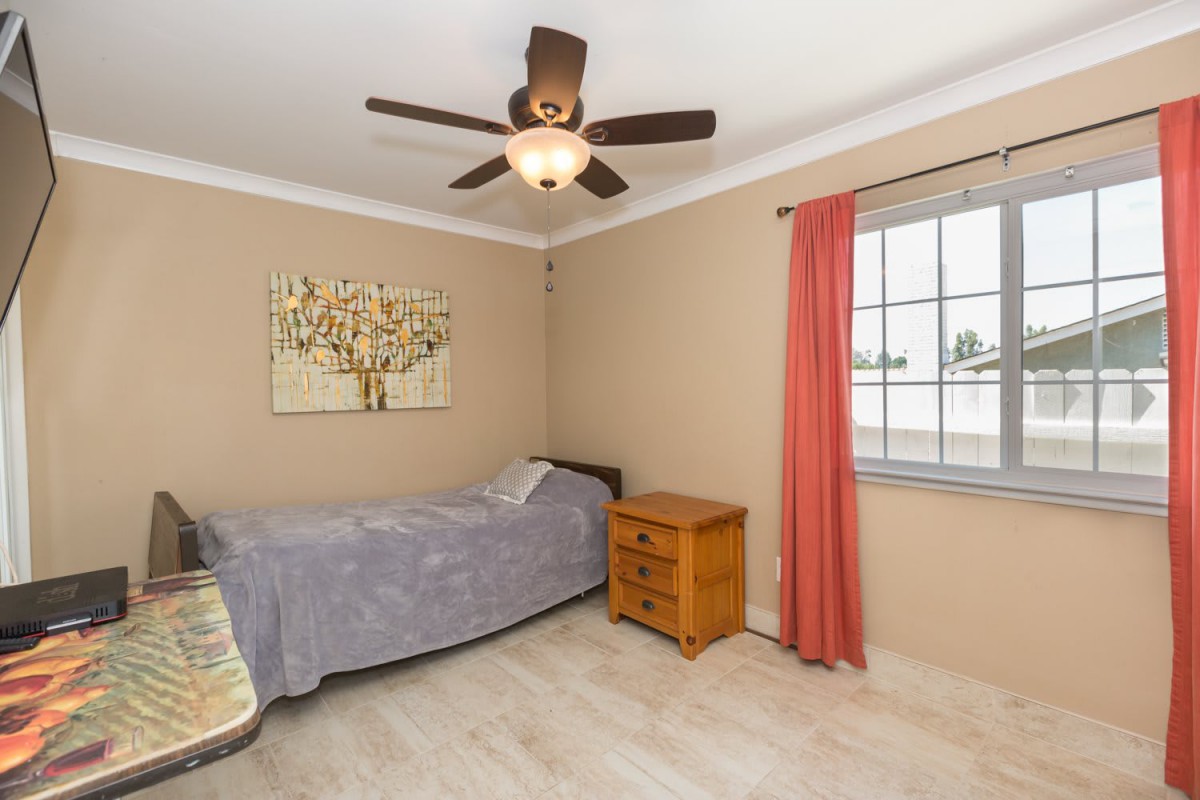 A cozy bedroom with a single bed, wooden nightstand, and a ceiling fan, featuring beige walls and red curtains.