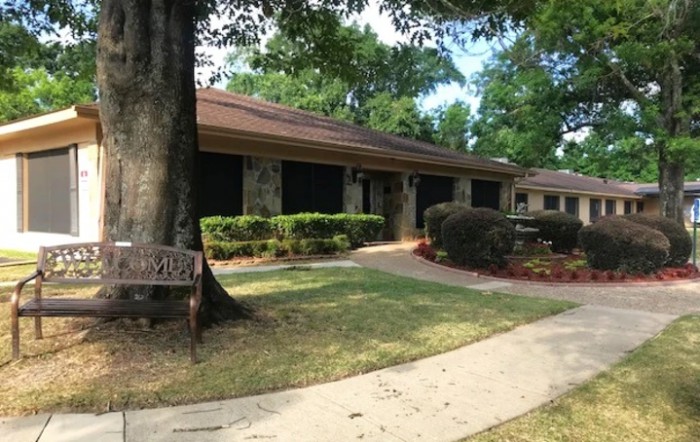 A one-story house surrounded by trees and shrubs, with a pathway leading to the entrance.