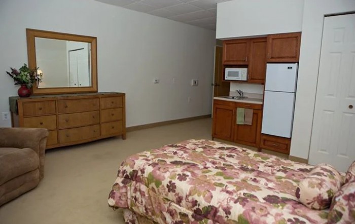 A cozy bedroom with a floral bedspread, wooden dresser with mirror, and a kitchenette featuring a refrigerator and microwave.