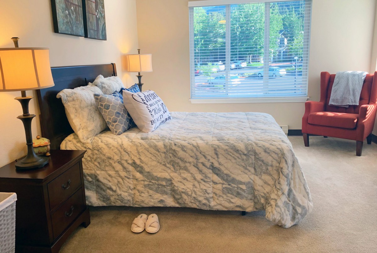 A cozy bedroom featuring a neatly made bed with patterned bedding, a bedside lamp, and a window letting in natural light.