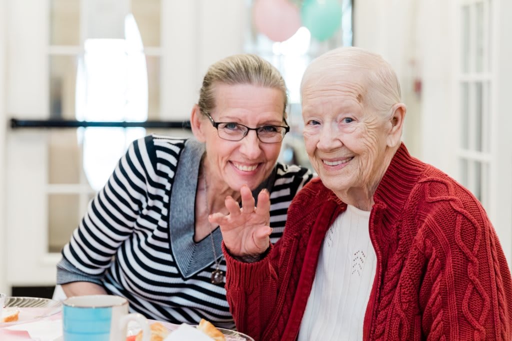 An elderly woman and a younger woman smiling and sitting together indoors.